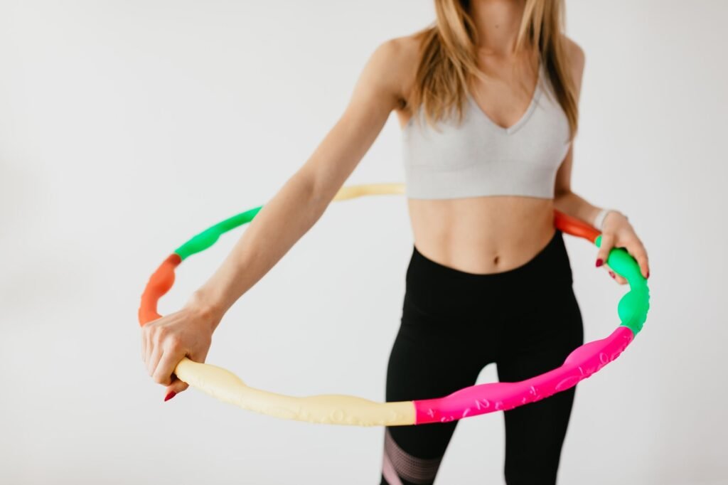 Crop faceless fit female in black leggings and gray sports bra standing with gymnastic hula hoop on gray background
