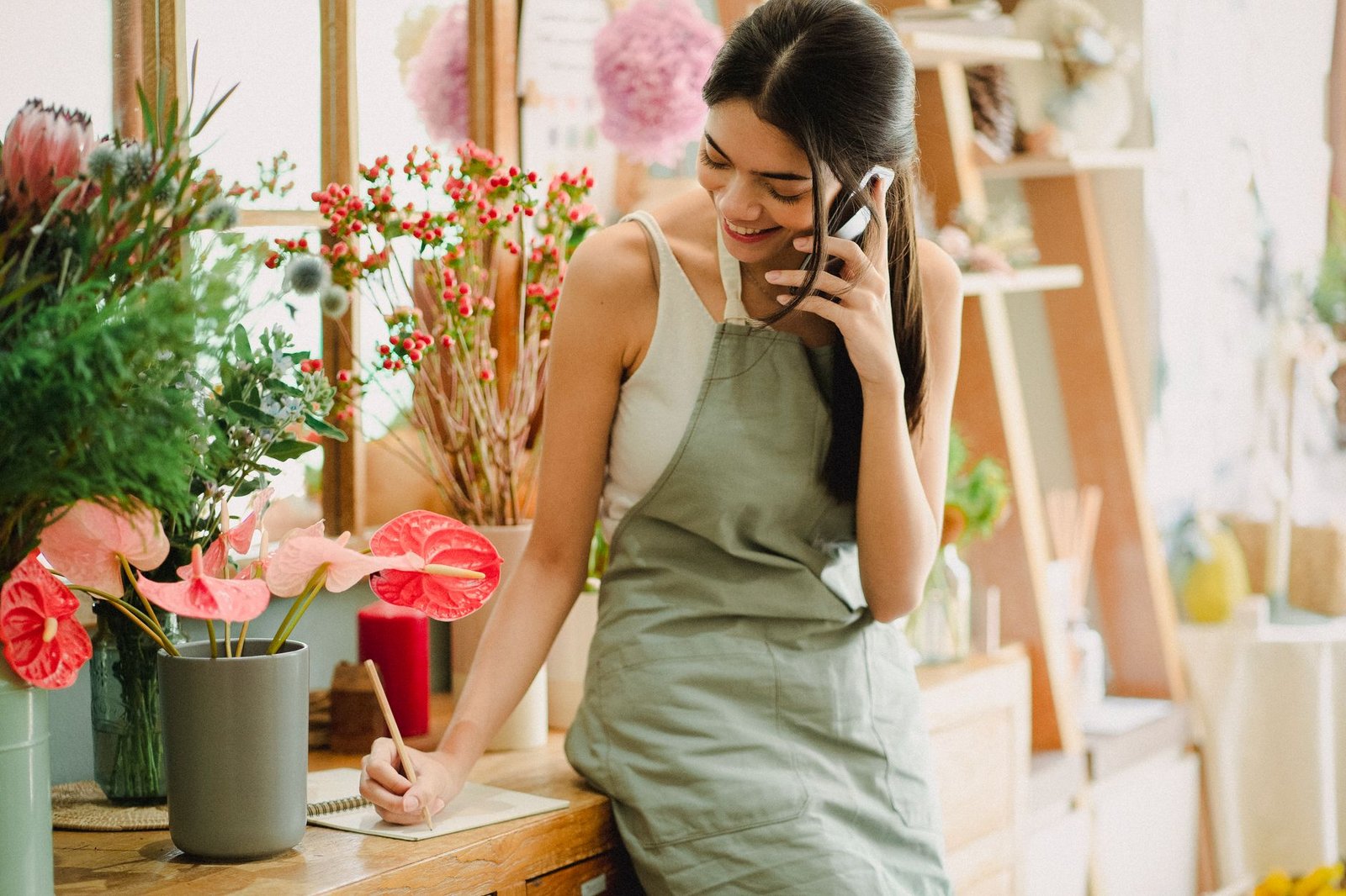 Smiling female florist in apron talking on mobile phone while standing near table and working in floral shop