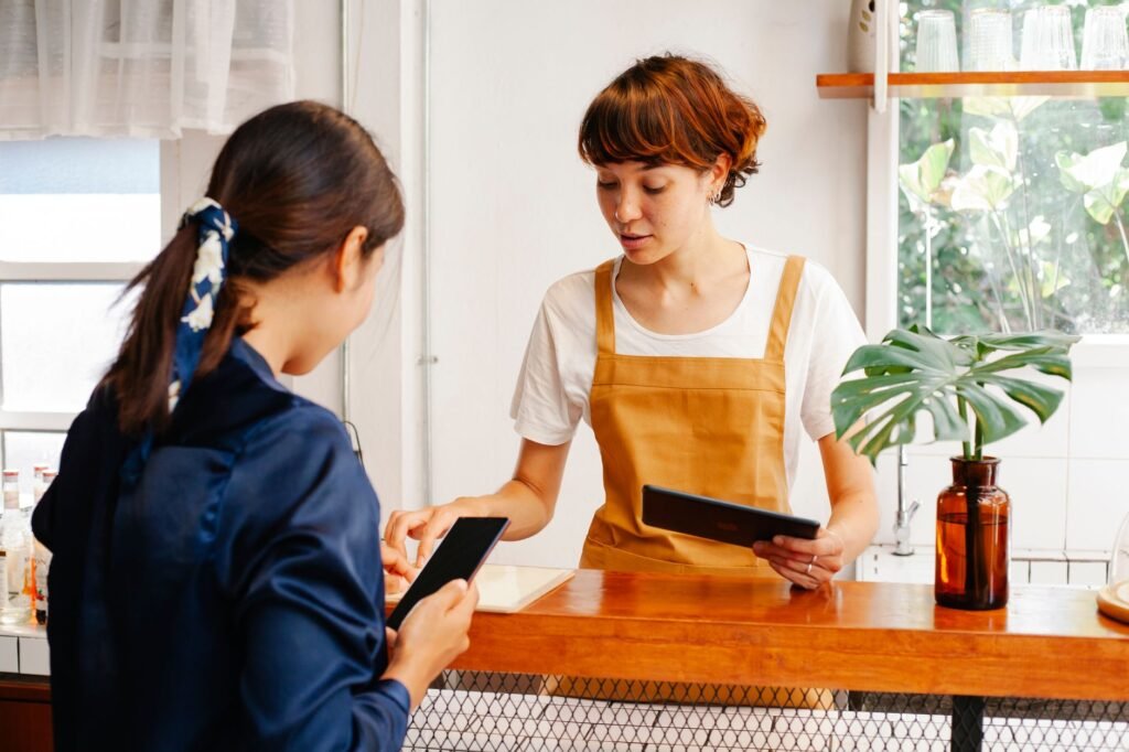 Unrecognizable female cafeteria worker with cellphone talking to young colleague with tablet in daylight