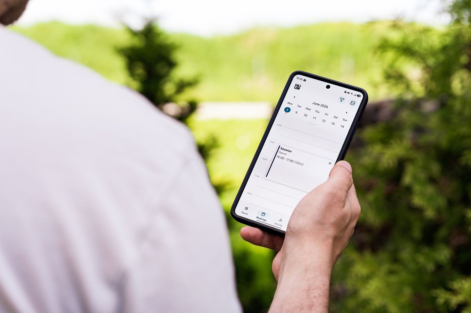 A person using a sauna booking app on a smartphone outdoors, surrounded by greenery.