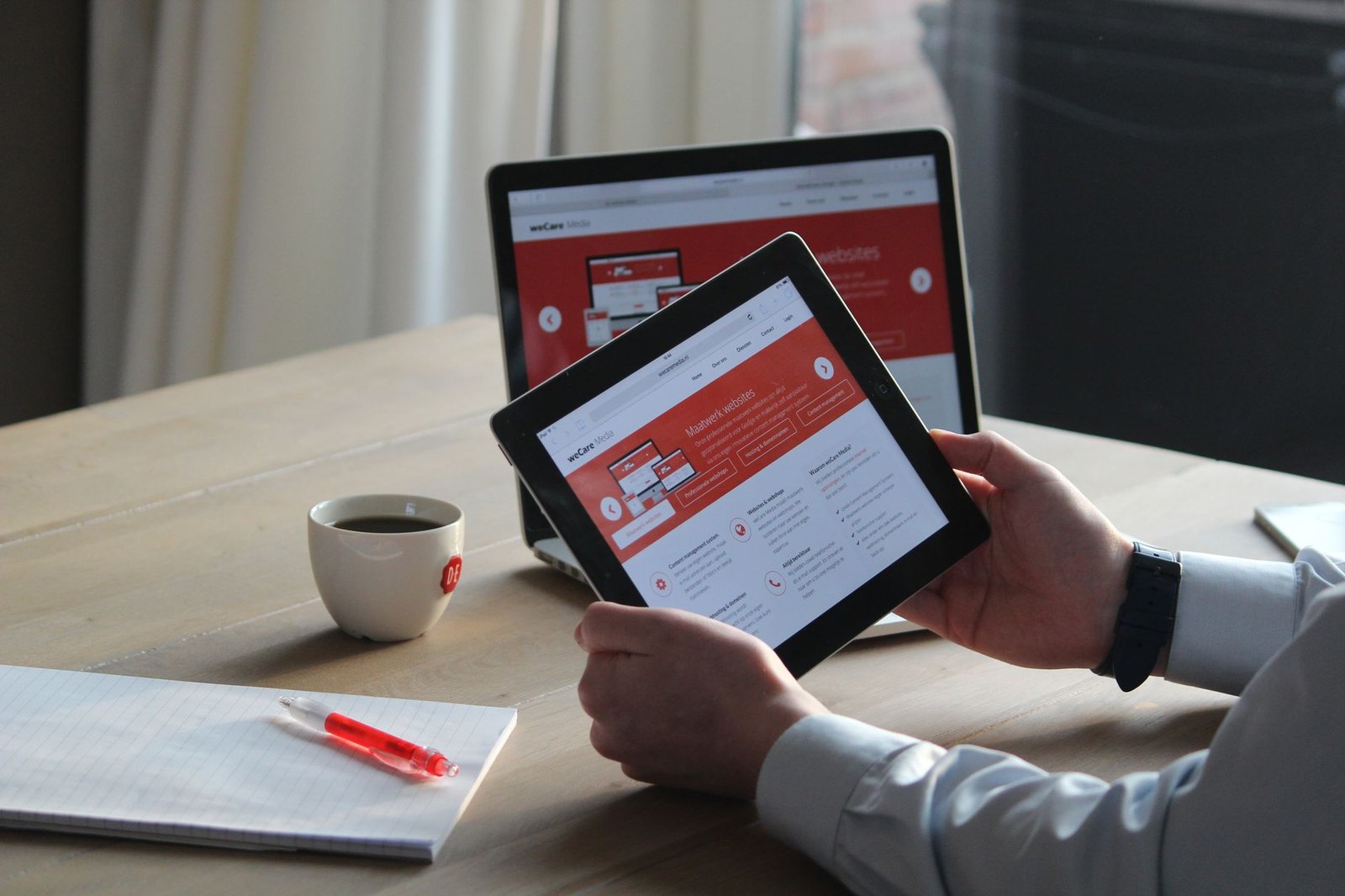 Business professional using a tablet and laptop with a hot drink, focusing on digital content review.