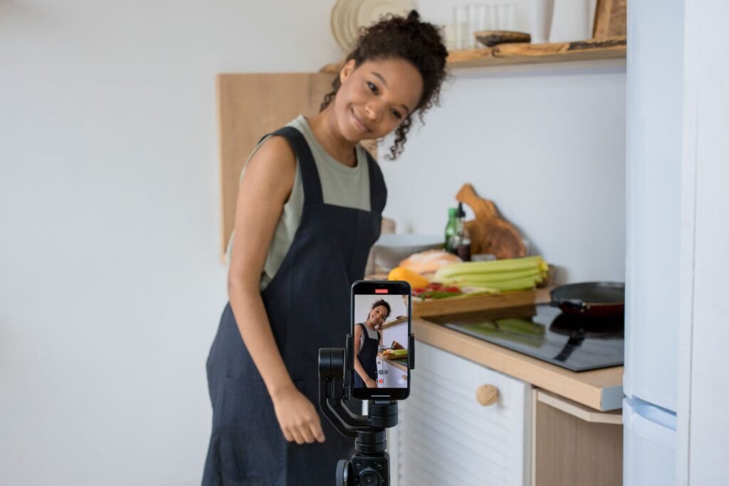 Smiling young woman films a cooking video at home using a smartphone in her kitchen.