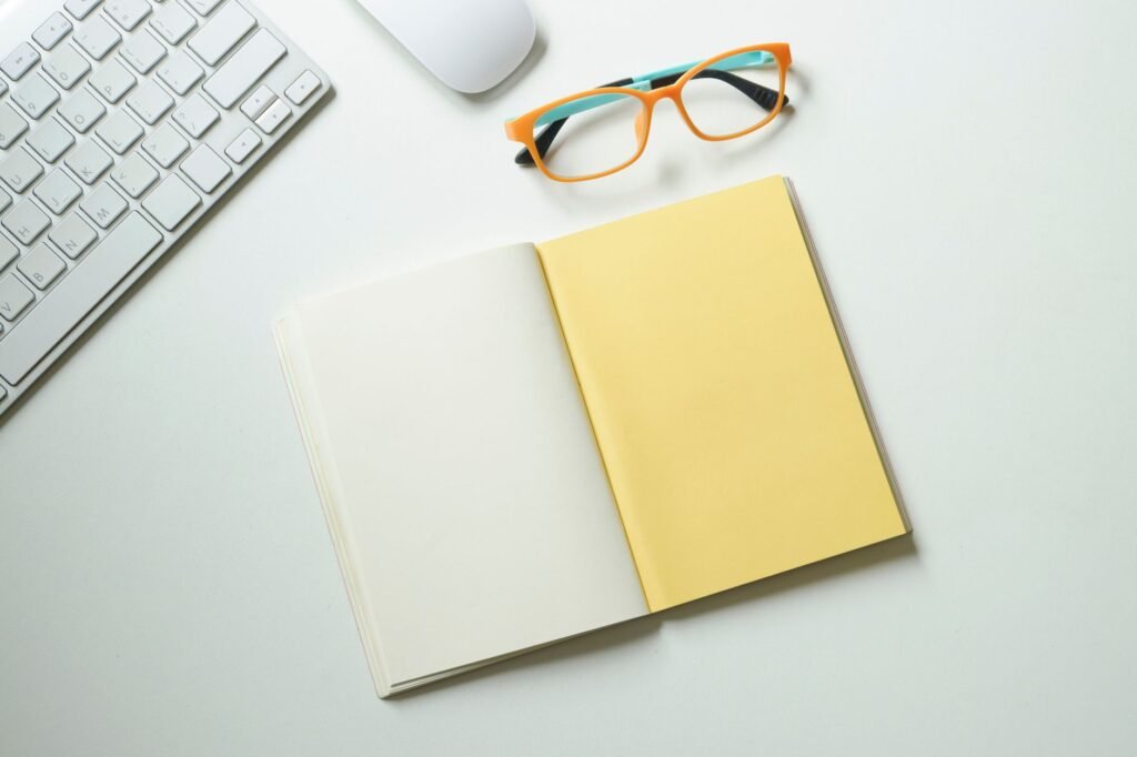 Top-down view of a clean workspace featuring a keyboard, mouse, stylish glasses, and an open notebook.