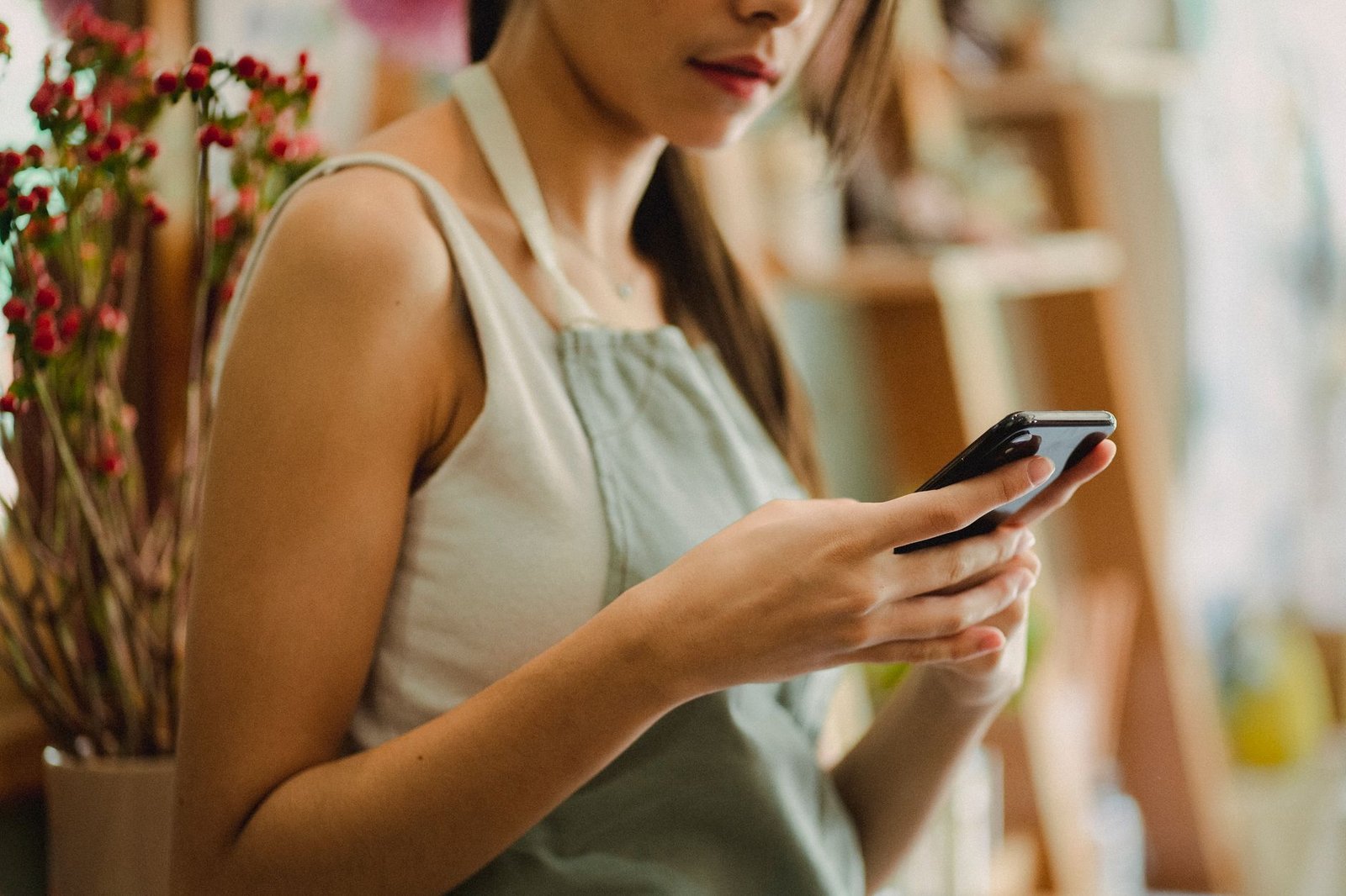 Crop anonymous florist in apron browsing mobile phone while working in cozy floral shop