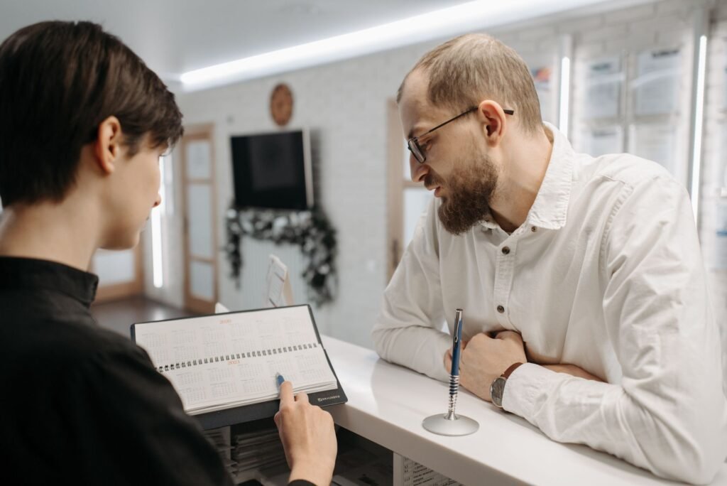 A receptionist and client converse over an appointment book at a clinic reception desk.