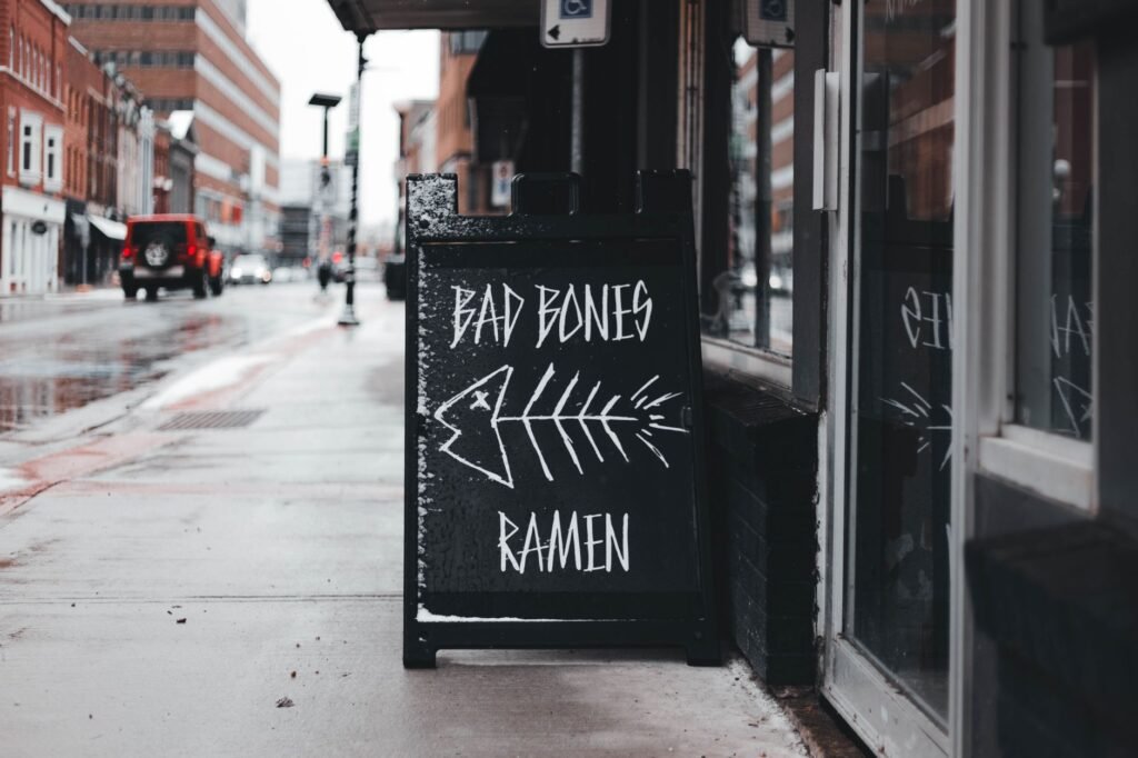 Moody street view showcasing a 'Bad Bones Ramen' chalkboard sign on a rainy day.